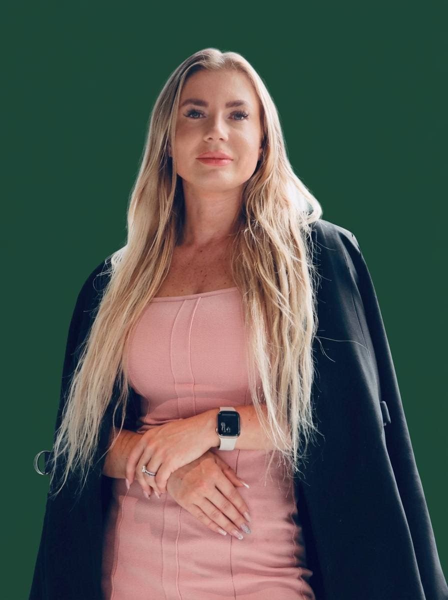 Portrait of Monika Flores Martin — blonde hair, light-pink bandeau dress under a dark jacket, hands clasped in front, on a deep green backdrop