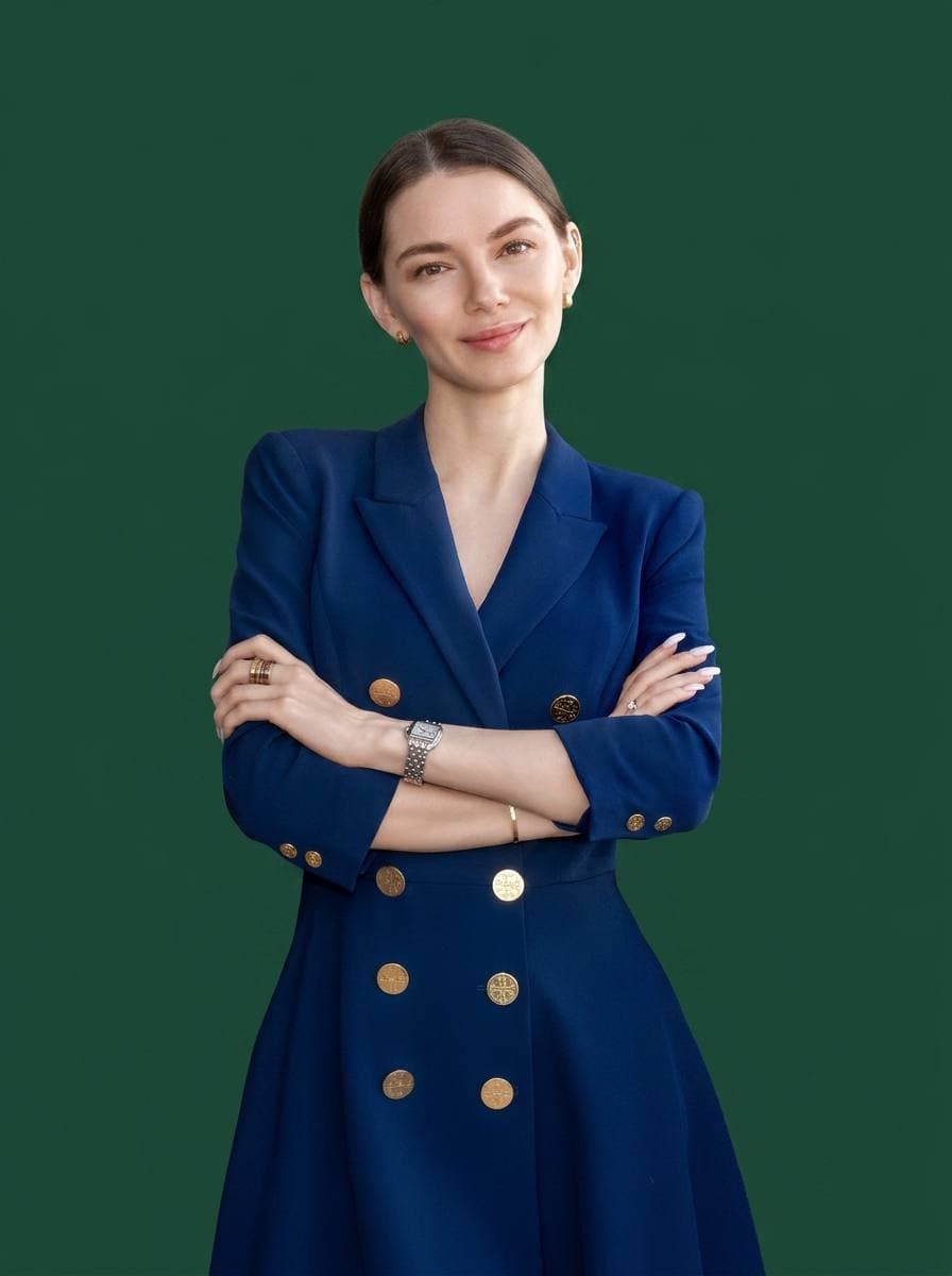 Portrait of Mariia Goncharova — brown hair pulled back, deep-blue double-breasted blazer with gold buttons, arms crossed, on a deep green backdrop