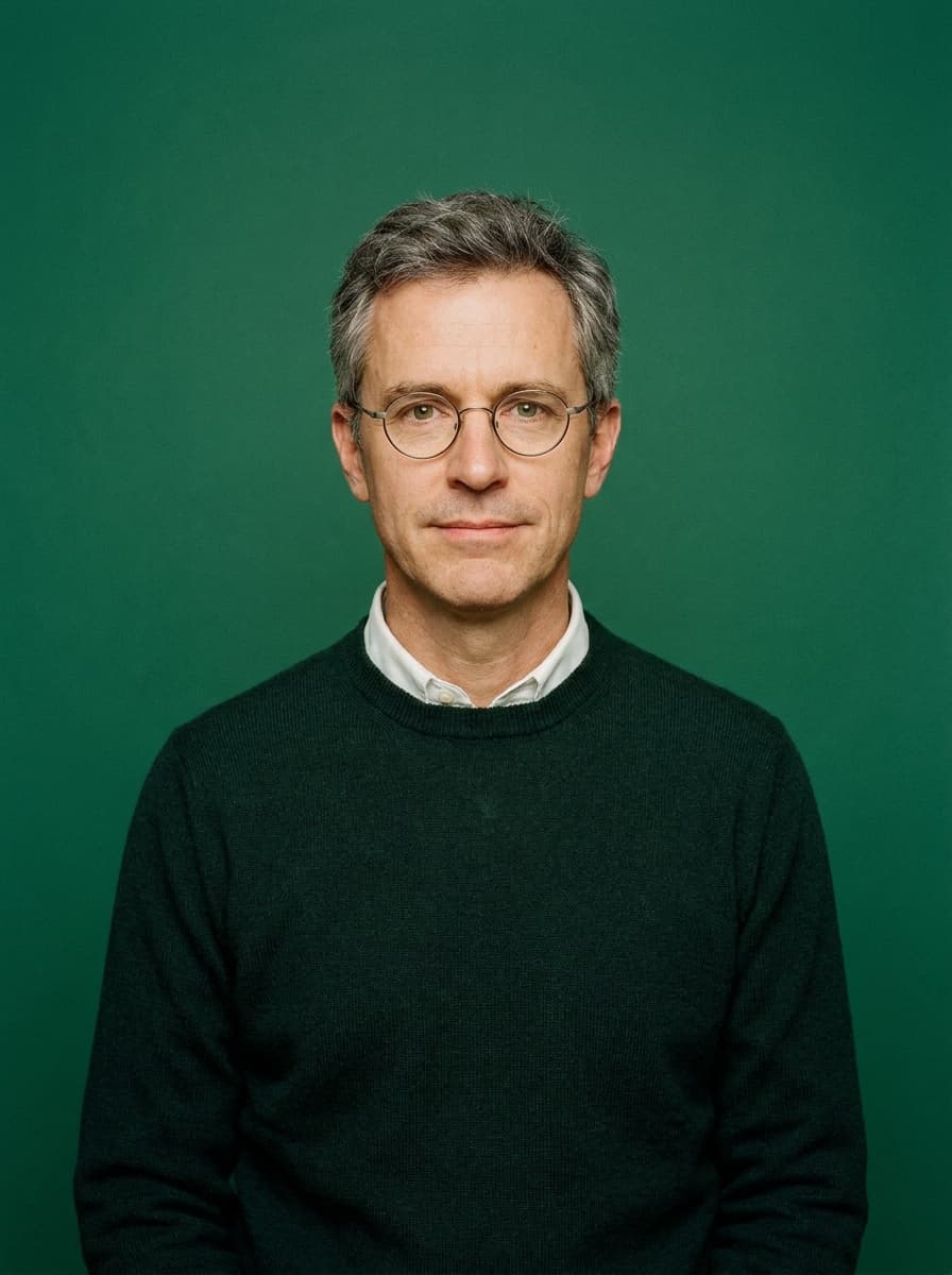 Portrait of Teodor Grech — round spectacles, dark-green sweater over a white shirt, on a deep green backdrop
